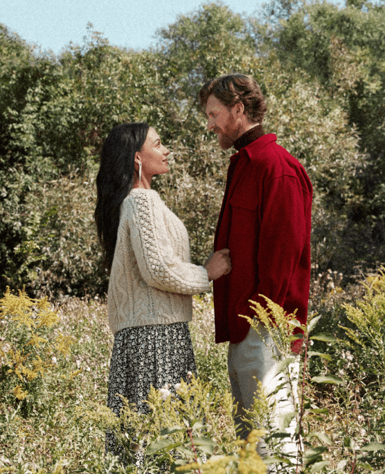 A man and a woman stand facing each other among trees and wildflowers.