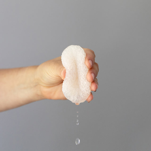 A hand squeezing the Konjac hydrating vegetable fibre sponge showing water droplets coming out of it against a grey background